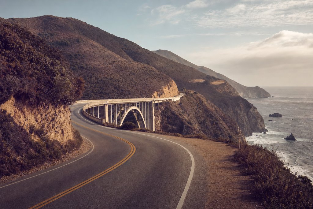 A two-lane highway on a cliff above the Pacific with a concrete arch bridge ahead. (AI-generated image)