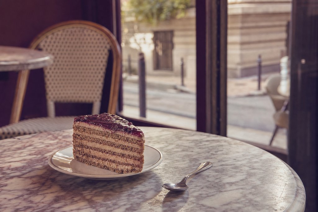 a slab of layered cake on a small plate beside a teaspoon on a marble table. (AI-generated image)