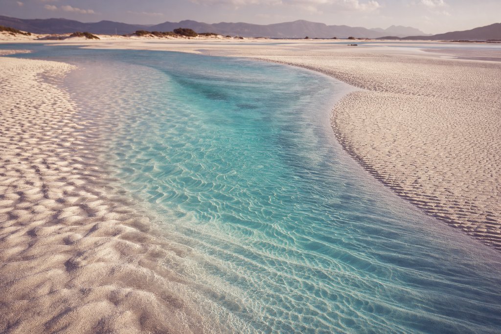 a shallow turquoise channel over rippled white sand seen from low altitude. (AI-generated image)