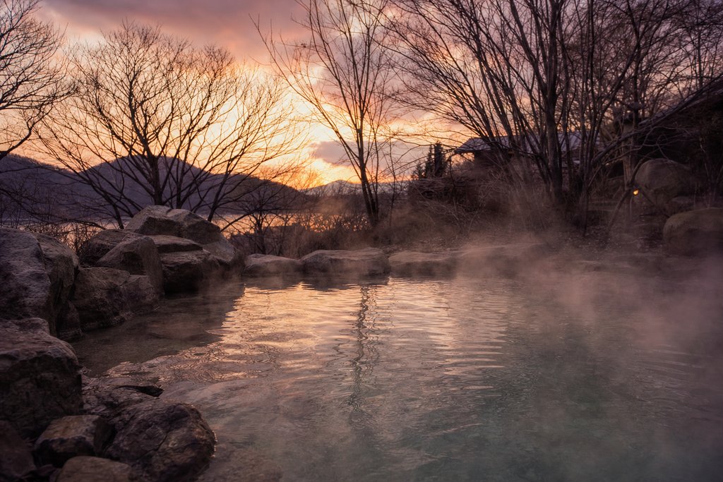 a steaming outdoor mineral pool ringed by rough stone and bare winter trees at dusk. (AI-generated image)