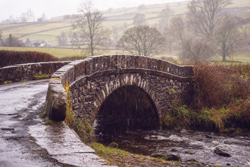 a wet stone bridge over a small river between hedgerows in soft rain. (AI-generated image)
