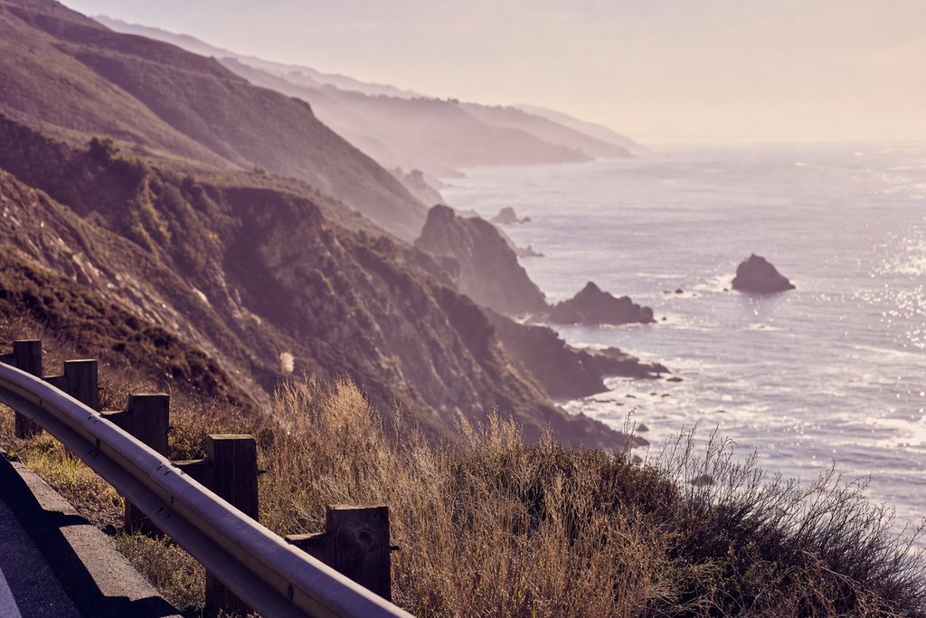 a coastal guardrail above a rocky Pacific cove in afternoon haze. (AI-generated image)
