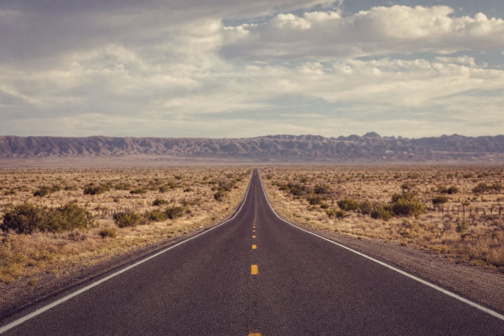 a long straight two-lane road across flat scrub toward a distant ridge. (AI-generated image)