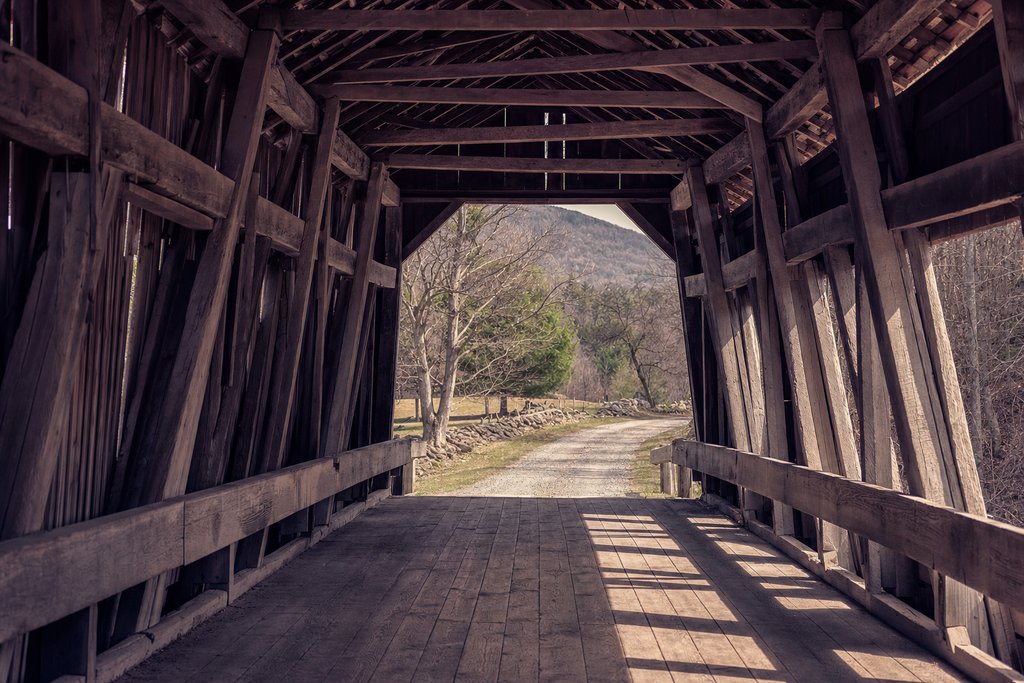 The inside of a timber covered bridge looking out through the far opening onto a gravel lane. (AI-generated image)