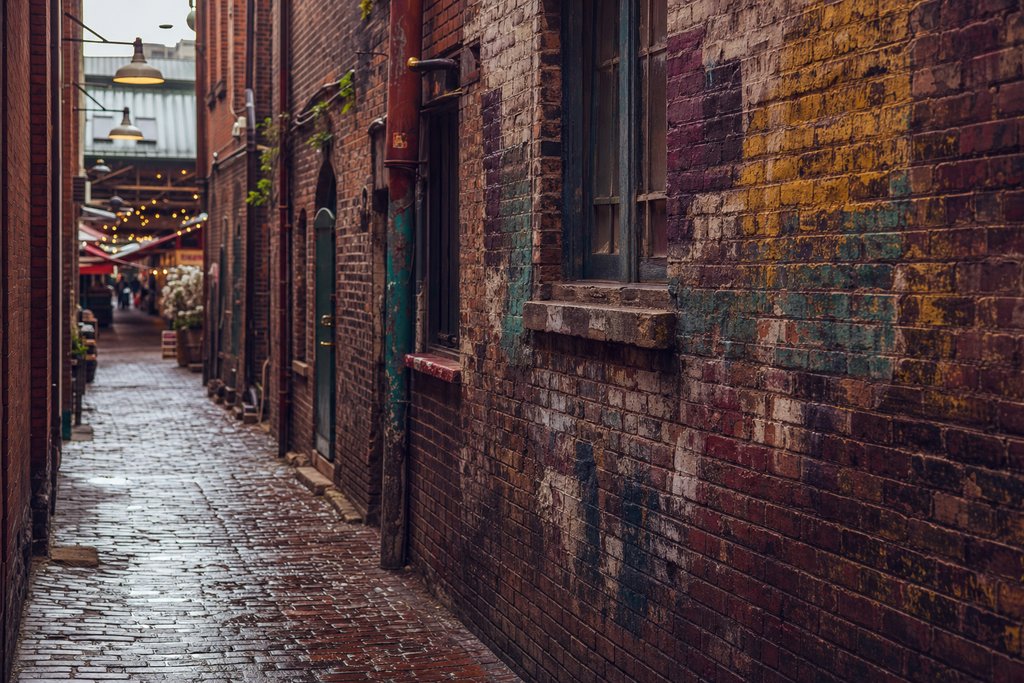 A narrow brick service alley below a public market, walls thickly encrusted with layered colour, wet cobbles. (AI-generated image)