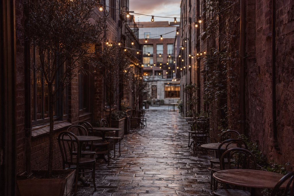 A narrow bluestone laneway at dusk strung with festoon lights above empty café chairs. (AI-generated image)