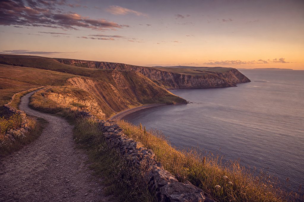 A clifftop coast path with a weathered timber signpost above a wide bay at golden hour. (AI-generated image)