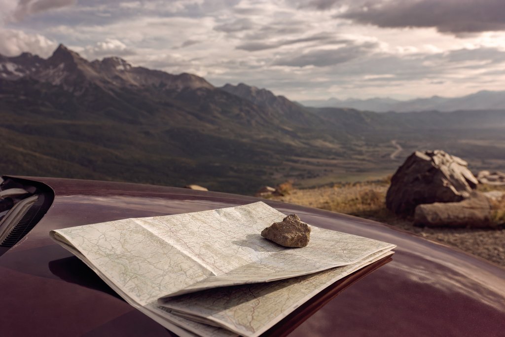A folded paper road map weighted with a stone on a car bonnet at a mountain lookout. (AI-generated image)
