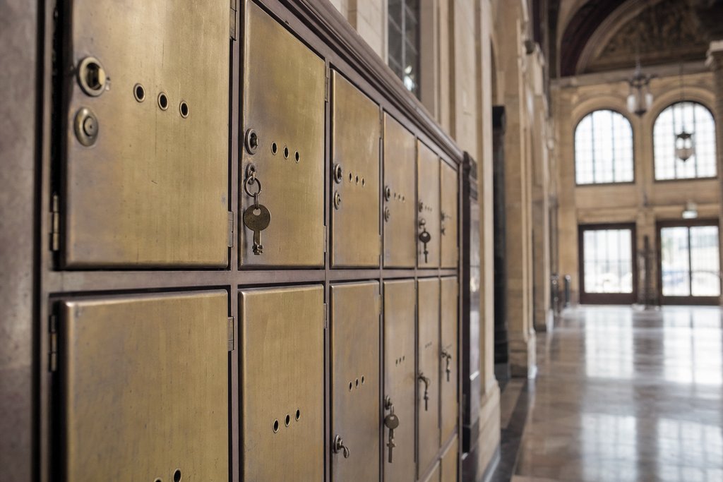 A row of brass left-luggage lockers in a railway station concourse, several keys still in the doors, cool daylight. (AI-generated image)