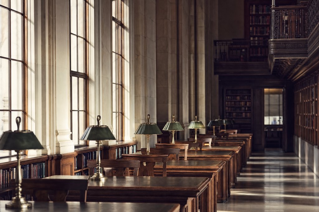 An empty library reading room with long timber tables and low green lamps, tall windows. (AI-generated image)