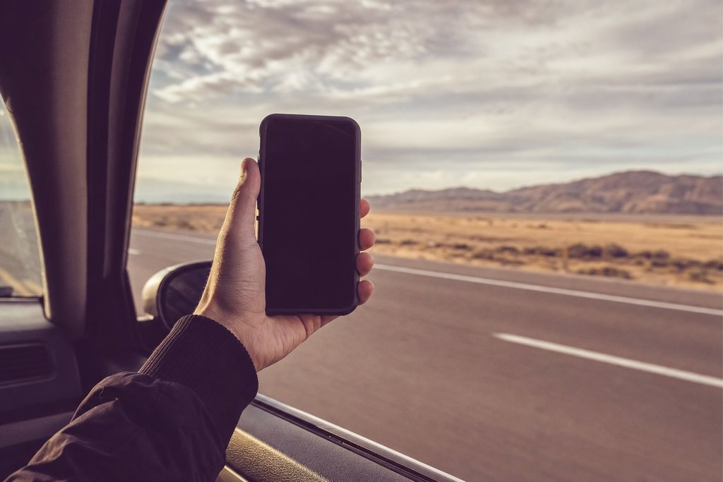 A hand holding a phone against a car window with open highway beyond, screen dark. (AI-generated image)