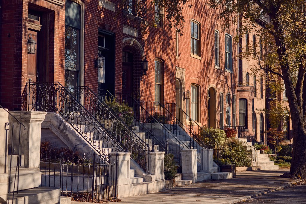 A block of brick rowhouses with white marble stoops in low sun, Baltimore. (AI-generated image)