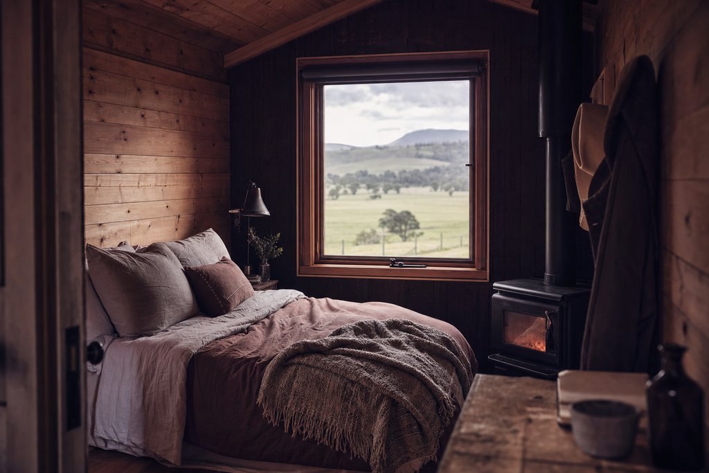 The interior of a tiny timber cabin with a bed, a small stove and a window over paddocks. (AI-generated image)