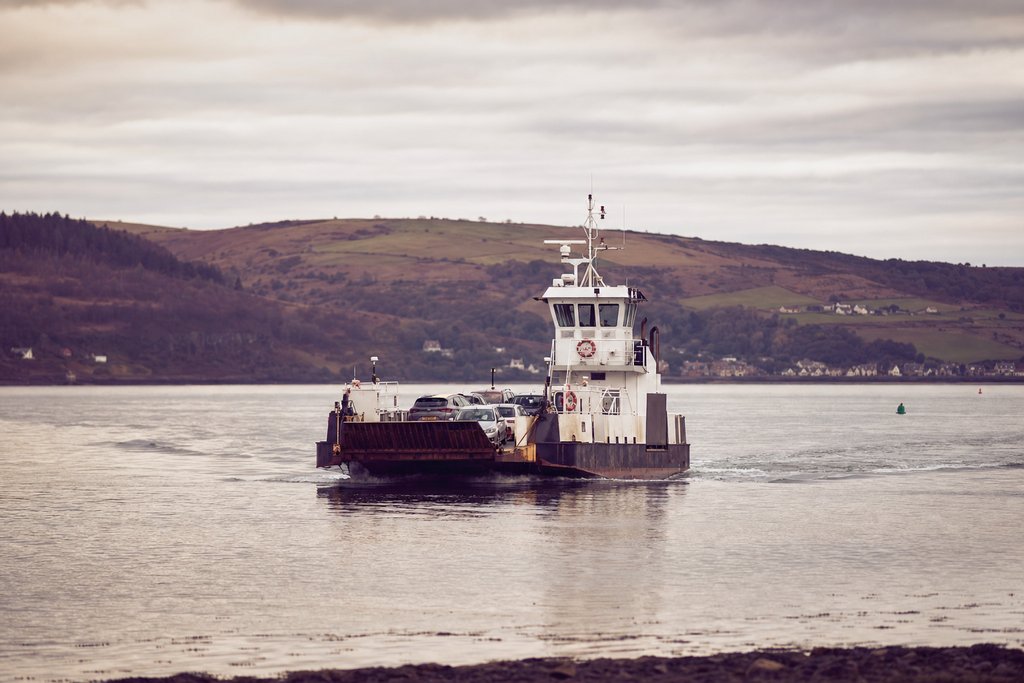 A small vehicle ferry crossing a wide estuary on flat grey water, wake trailing behind. (AI-generated image)