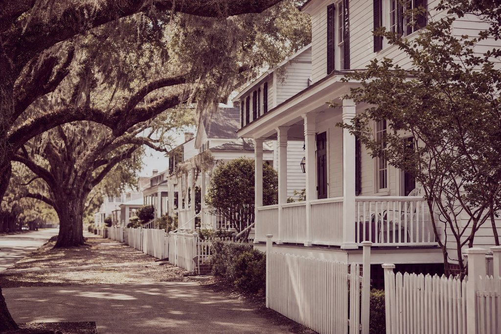 A street of white clapboard cottages with picket fences under old live oaks. (AI-generated image)