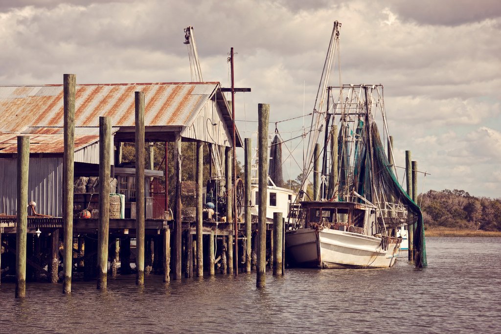 A working shrimp dock of wooden pilings and a tin-roofed fish house on a tidal river. (AI-generated image)