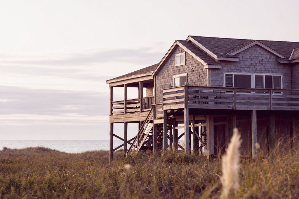 A weathered timber beach house raised on pilings above dune grass, Outer Banks, flat morning light. (AI-generated image)