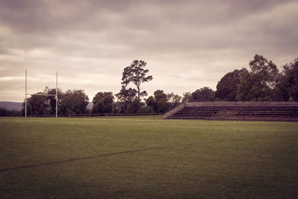 An empty grass playing field with goalposts and a low timber bleacher under overcast sky. (AI-generated image)