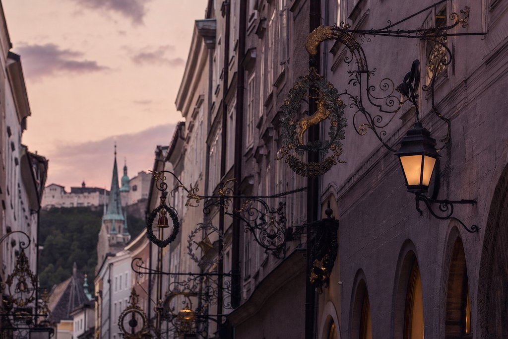A narrow old-town lane with wrought-iron guild brackets at dusk, Salzburg. (AI-generated image)