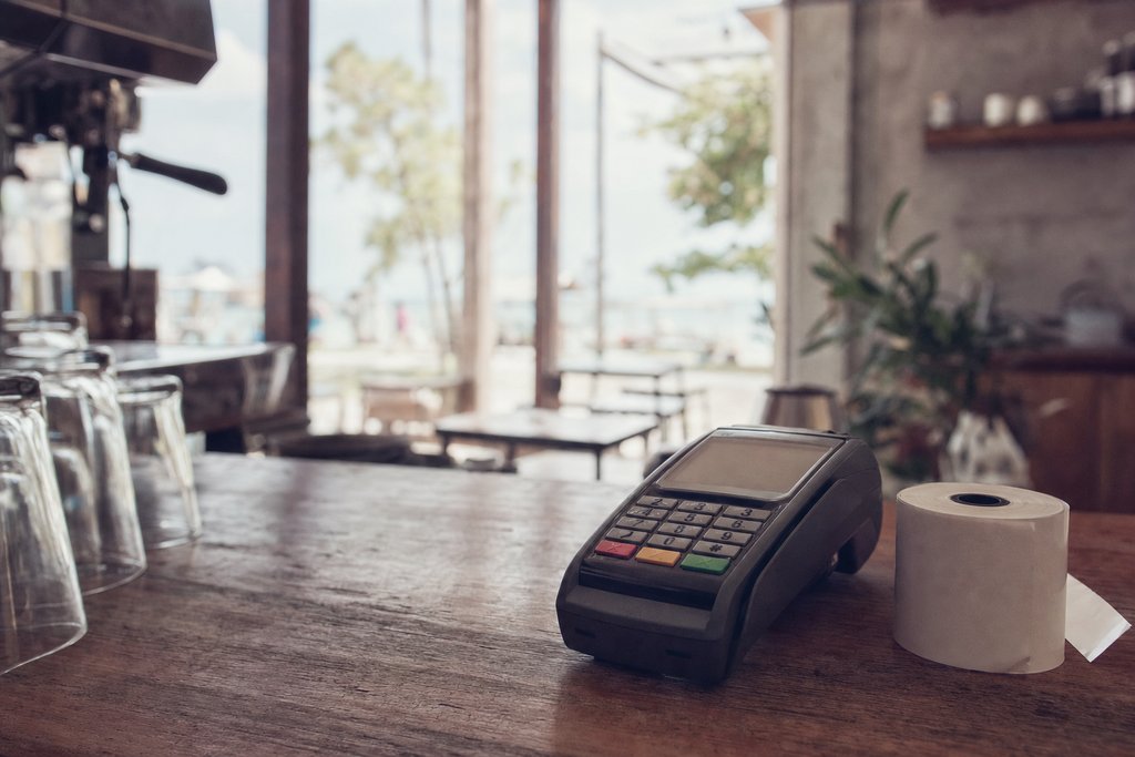 A card terminal on a timber café bar beside a receipt spool, shallow focus. (AI-generated image)