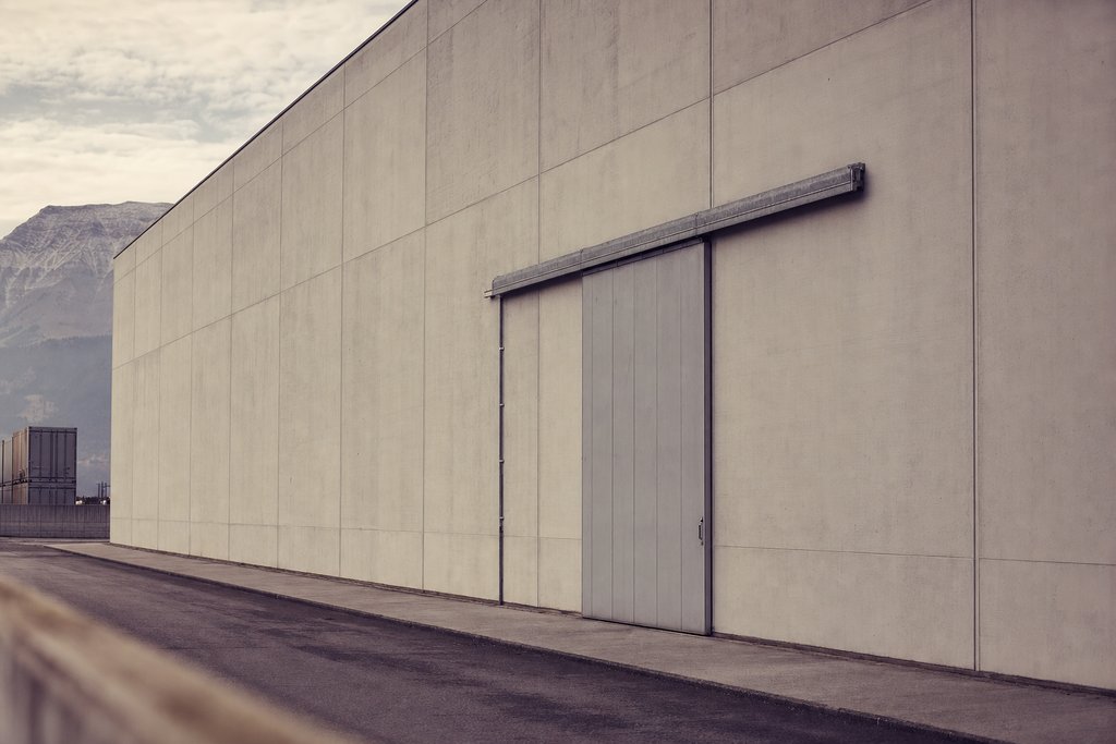 A vast windowless pale grey warehouse wall with a tall sliding service door. (AI-generated image)