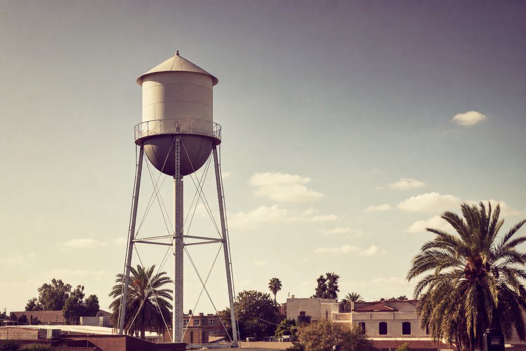 A steel-legged water tower with a blank tank standing over low buildings and palms. (AI-generated image)